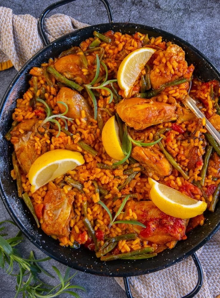 Paella with chicken, green beans, and lemon slices in a pan on a textured background.