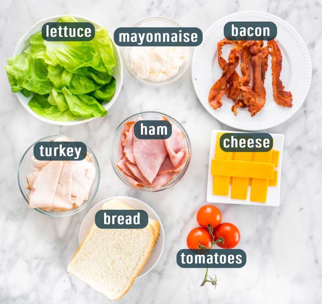 Sandwich ingredients: lettuce, mayo, bacon, turkey, ham, cheese, bread, tomatoes on a white marble surface.