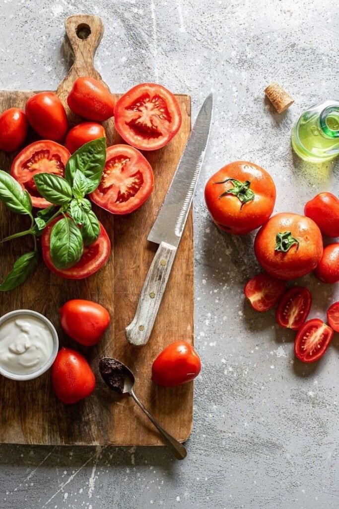 Fresh tomatoes and basil on a wooden board with knife, surrounded by olive oil, cream, and spices on a gray surface.