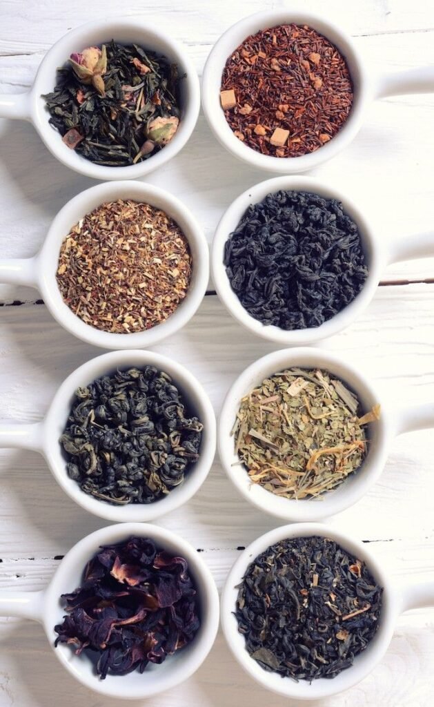 Assorted tea leaves in white cups on a wooden surface, showcasing green, black, and herbal tea varieties.