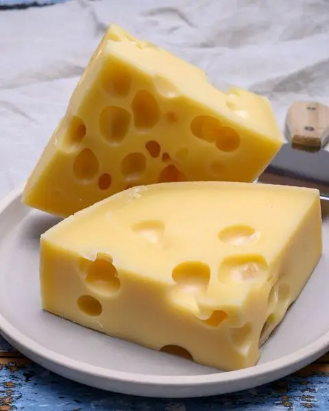 Two wedges of Swiss cheese with characteristic holes on a plate, knife in the background.