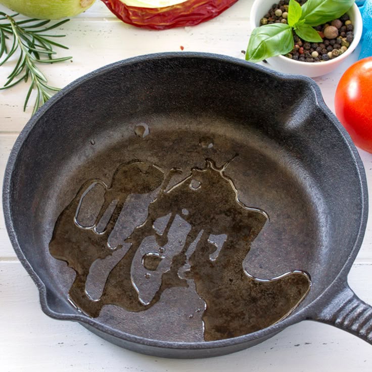 Cast iron pan with oil surrounded by herbs, spices, and vegetables on a white table. Cooking preparation scene.