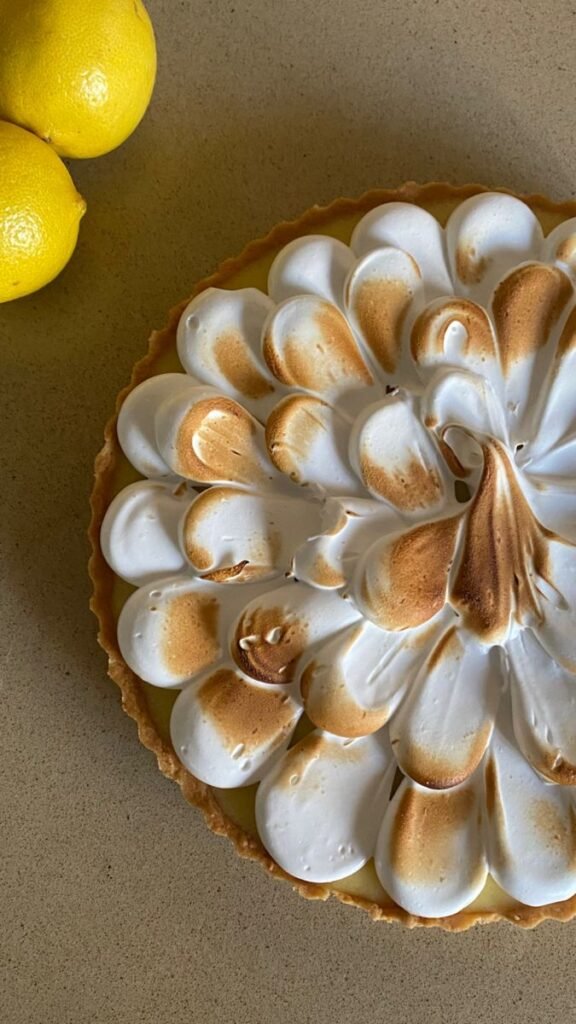 Lemon meringue pie with toasted peaks next to fresh lemons on a countertop.