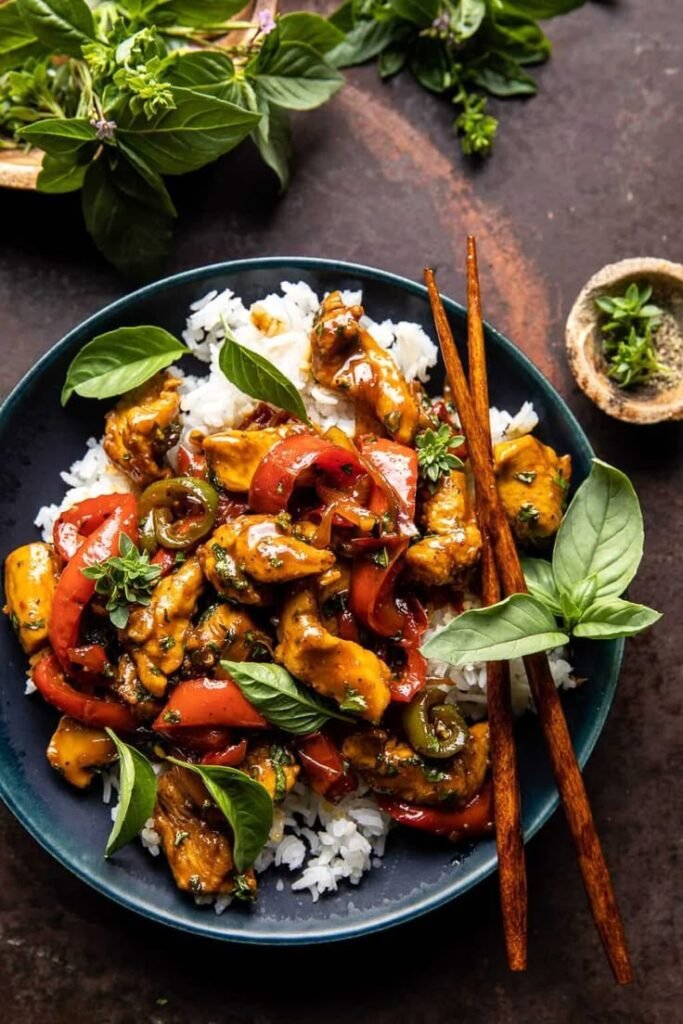 Stir-fried chicken with bell peppers and herbs on rice, served with chopsticks on a dark plate.