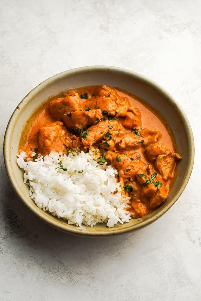 Bowl of creamy chicken curry with white rice garnish on a light background.