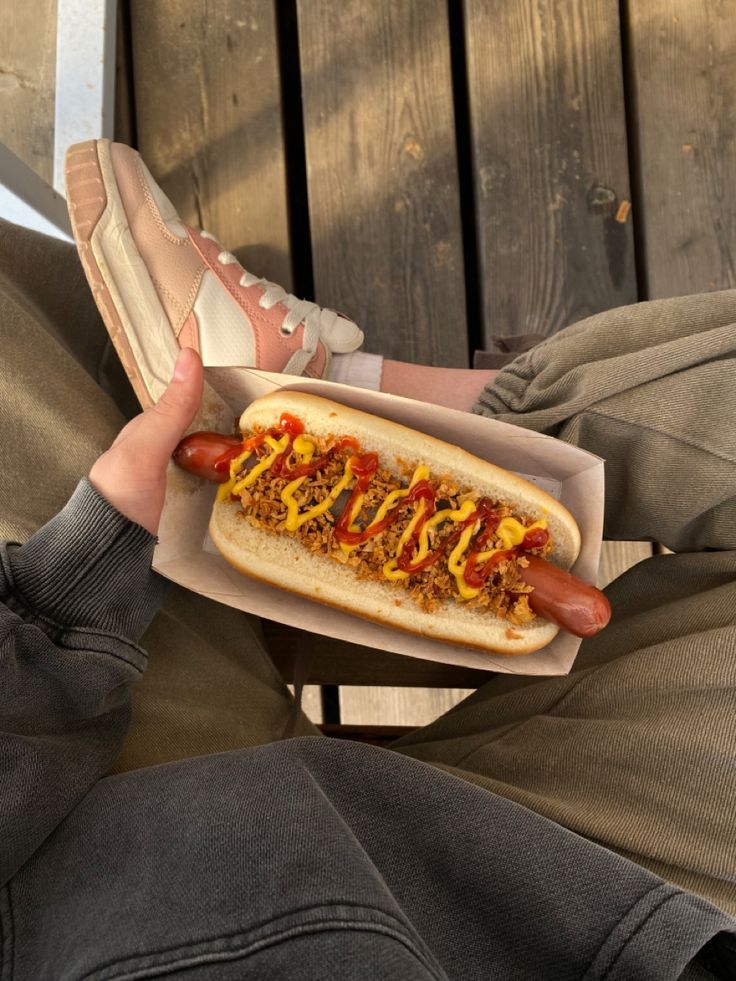 Person holding a chili cheese hot dog with mustard and ketchup, wearing casual attire on a wooden background.
