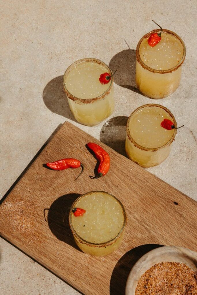 Spicy margaritas with chili garnish on a wooden board, sunlit summer cocktail setting.