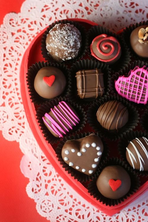 A heart-shaped box of assorted valentine chocolates with decorative designs on a lace and red background.