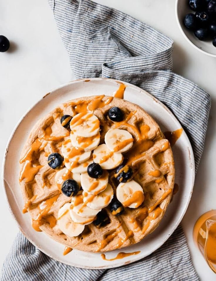 Waffle topped with banana slices, blueberries, and caramel sauce on a plate with a striped cloth background.