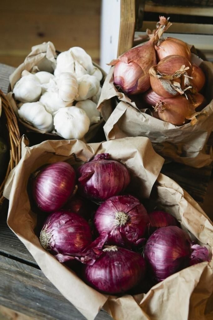 Baskets of fresh garlic, red onions, and shallots on a rustic wooden table.