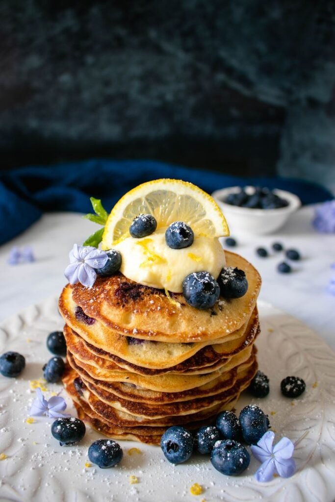 Stack of blueberry pancakes topped with whipped cream, lemon slice, and blueberries on a plate.