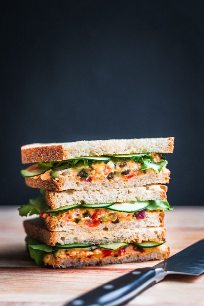 Stacked gourmet sandwiches with veggies and spread on rye bread against a dark background.