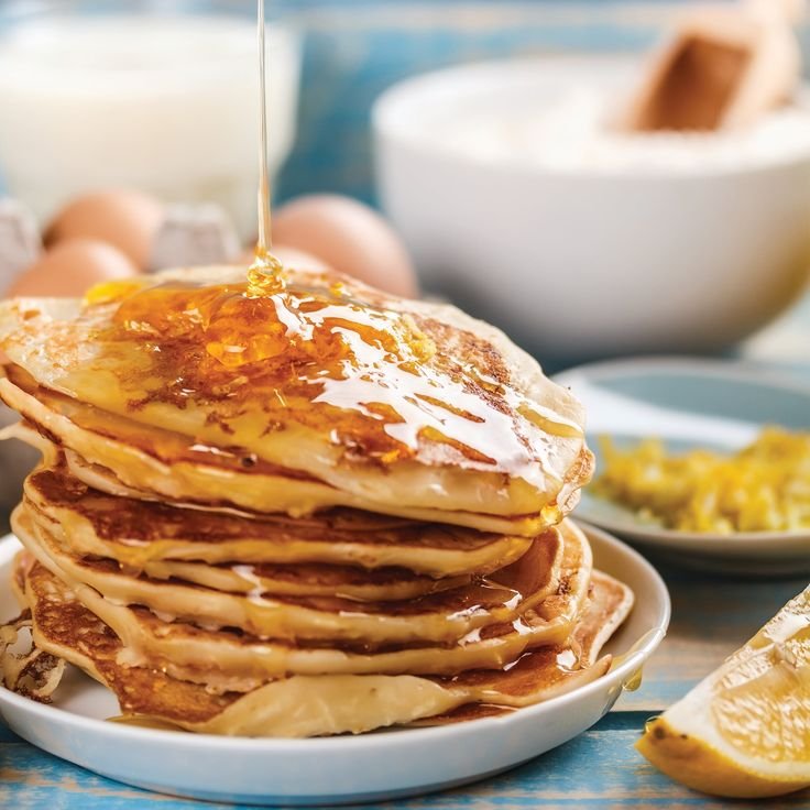 Stack of pancakes drizzled with honey and surrounded by ingredients on a table.