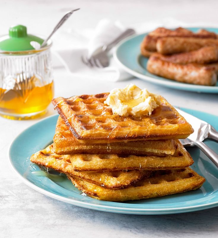 Stack of golden waffles with butter and syrup on a blue plate, accompanied by breakfast sausages and honey jar.