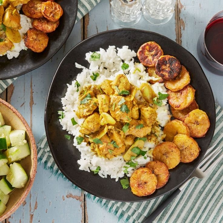 Plate of chicken curry with rice and fried plantains, served beside a cucumber salad.