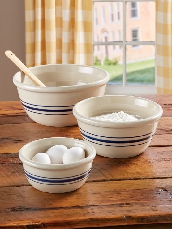 Three ceramic bowls with blue stripes on a wooden table, containing eggs, flour, and a wooden spoon by a window.