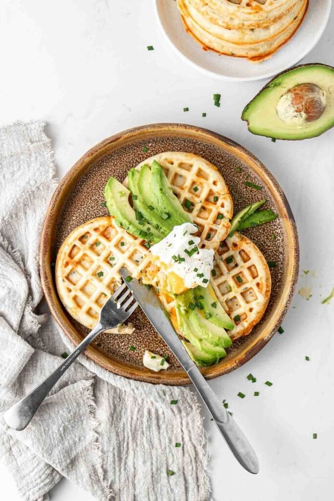 Savory waffles topped with avocado slices and a poached egg on a ceramic plate, garnished with chives.