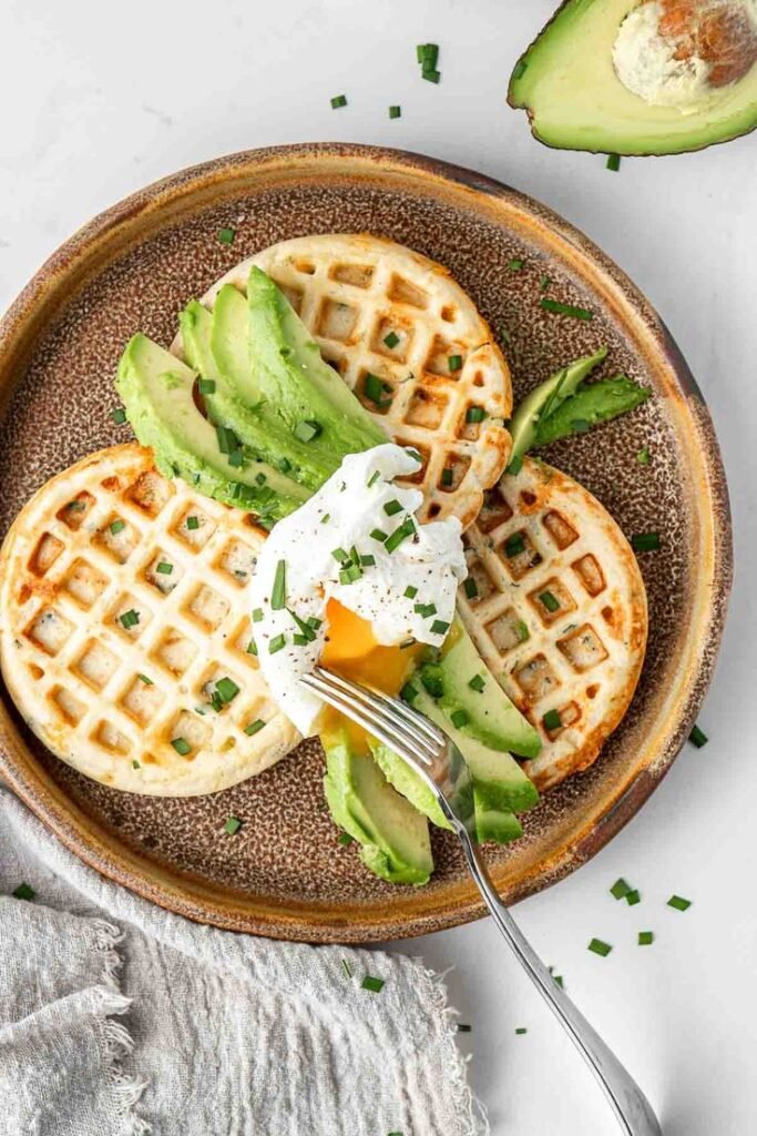 Avocado waffles topped with poached egg and chives served on a rustic plate.