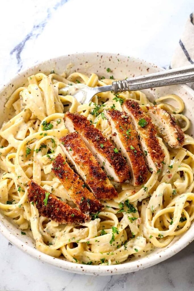 Creamy fettuccine Alfredo topped with crispy sliced chicken and fresh parsley in a bowl with a fork.