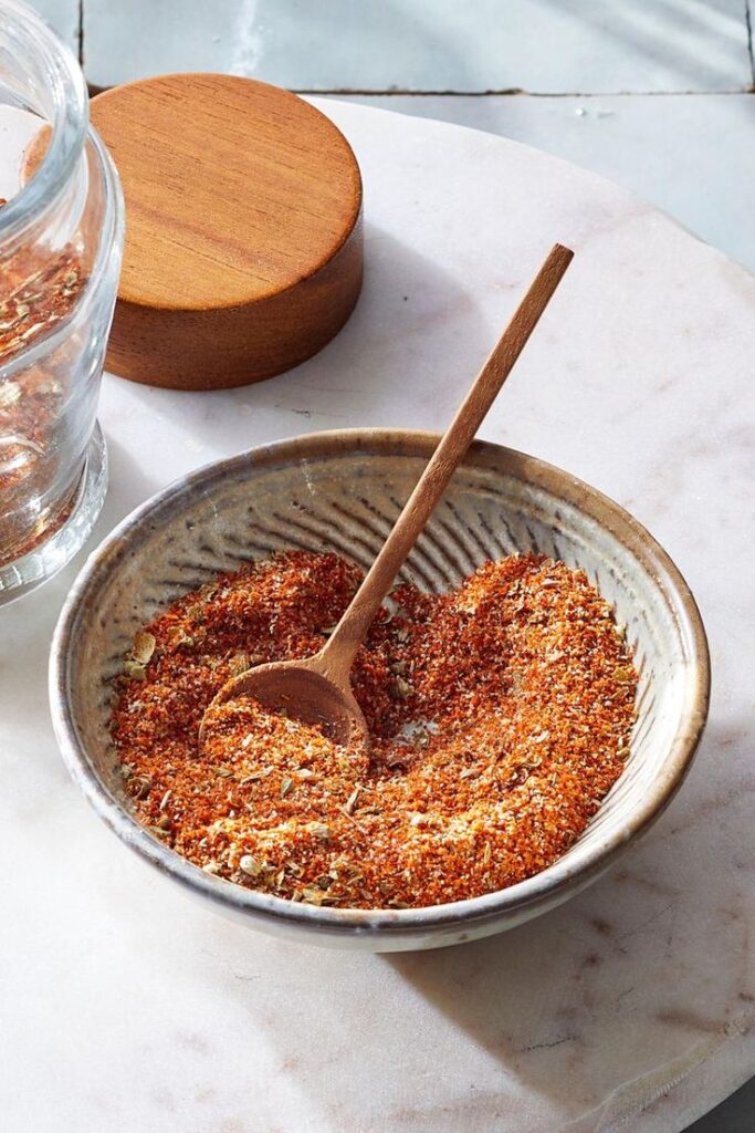 Ceramic bowl with spice blend and wooden spoon on marble surface, next to a glass jar with a wooden lid.
