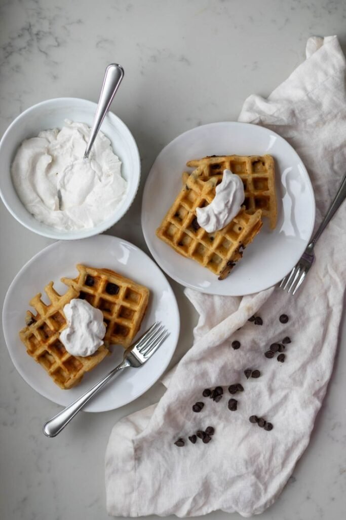 Waffles topped with whipped cream on plates, accompanied by bowls of cream and scattered chocolate chips.