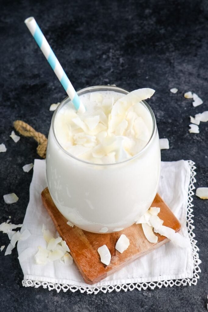 Creamy coconut smoothie with a striped straw on a wooden board, topped with coconut flakes. Refreshing and delicious.