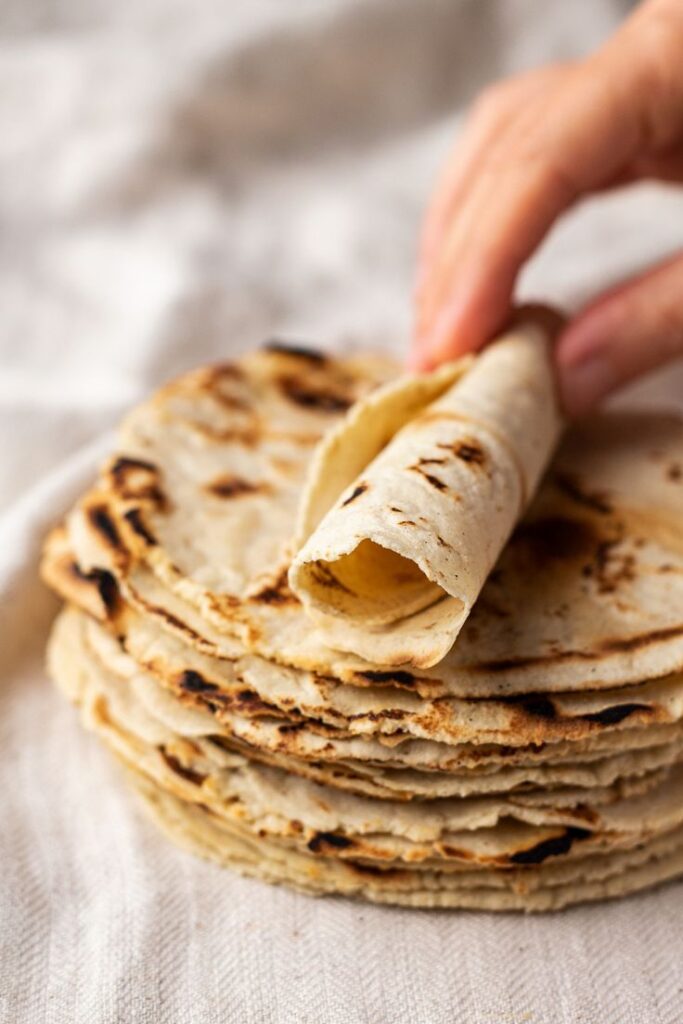 Stack of freshly made corn tortillas, with hand lifting one on a white cloth background.