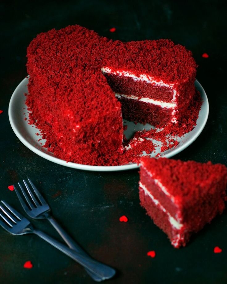 Heart-shaped red velvet cake with a slice cut, white frosting layers, and two forks on a dark background.