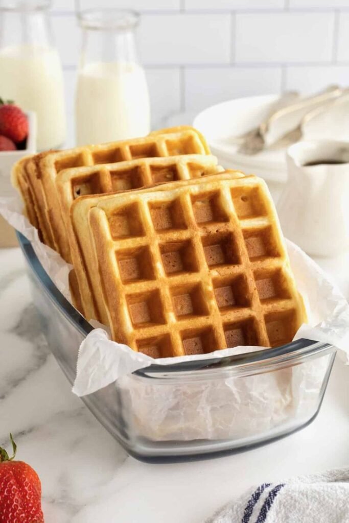 Golden waffles in a glass dish on a countertop, perfect for breakfast or brunch with fresh milk and strawberries.