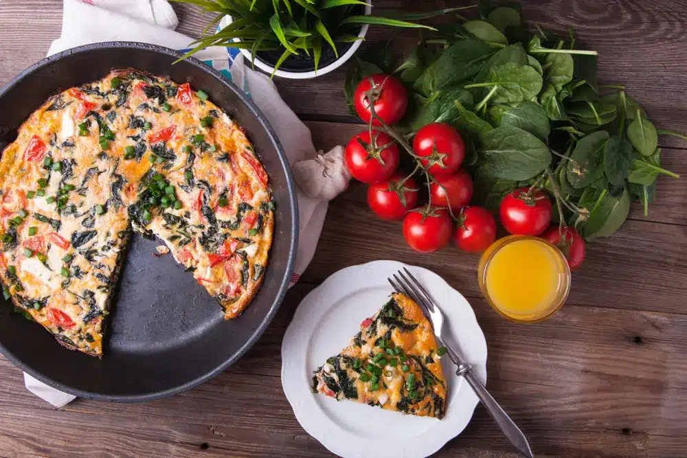 Vegetable frittata in a skillet with fresh tomatoes, spinach, and a glass of orange juice on a wooden table.