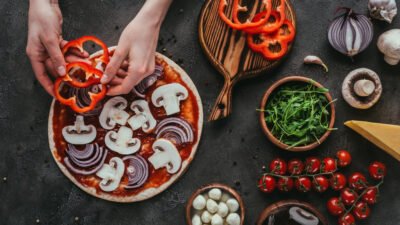 Hands adding red peppers to a pizza base with onions and mushrooms, surrounded by fresh ingredients.