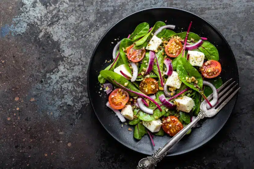 Fresh spinach salad with feta, cherry tomatoes, and red onion on a black plate with a fork, top view.
