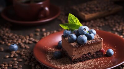 Chocolate mousse cake topped with blueberries and mint on a brown plate, surrounded by coffee beans.