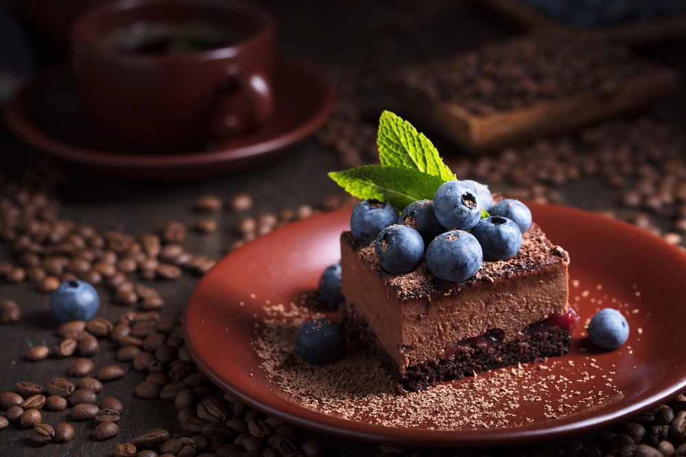 Chocolate mousse cake topped with blueberries and mint on a brown plate, surrounded by coffee beans.