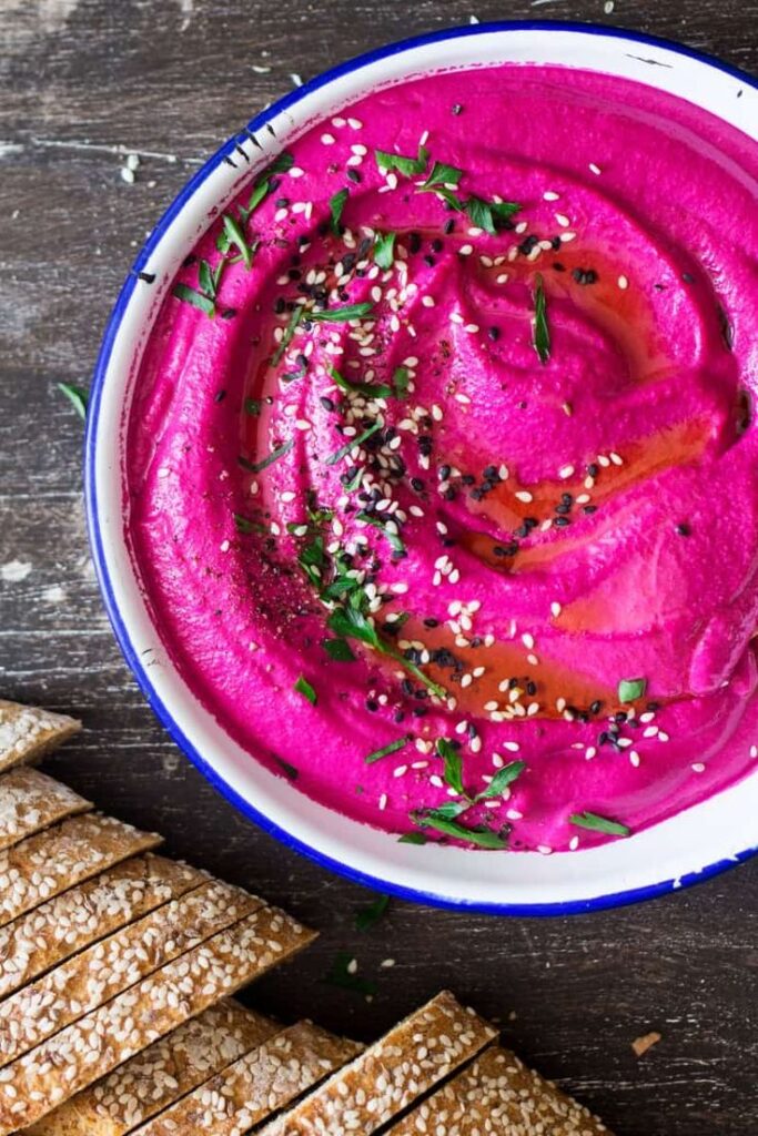 Vibrant pink beet hummus garnished with seeds and herbs, served with sesame seed crackers on a rustic wooden table.