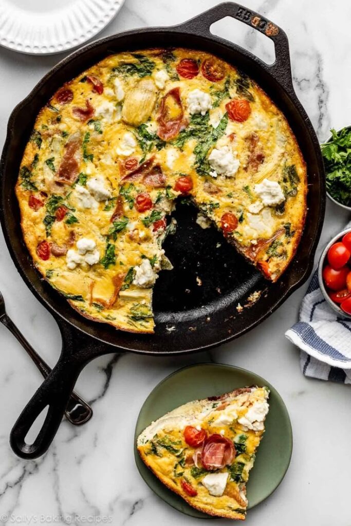 Delicious homemade frittata with cherry tomatoes, goat cheese, and herbs in a cast iron skillet, slice served on a plate.