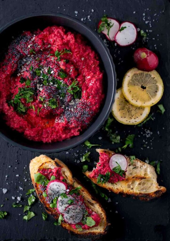 Beetroot hummus in a bowl with toasted bread, garnished with radishes, herbs, and lemon slices on a dark surface.
