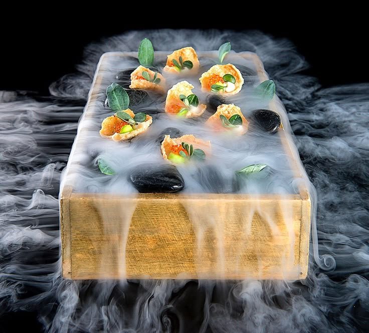 Elegant gourmet appetizers in a wooden box with dramatic smoke, garnished with herbs and stone accents.