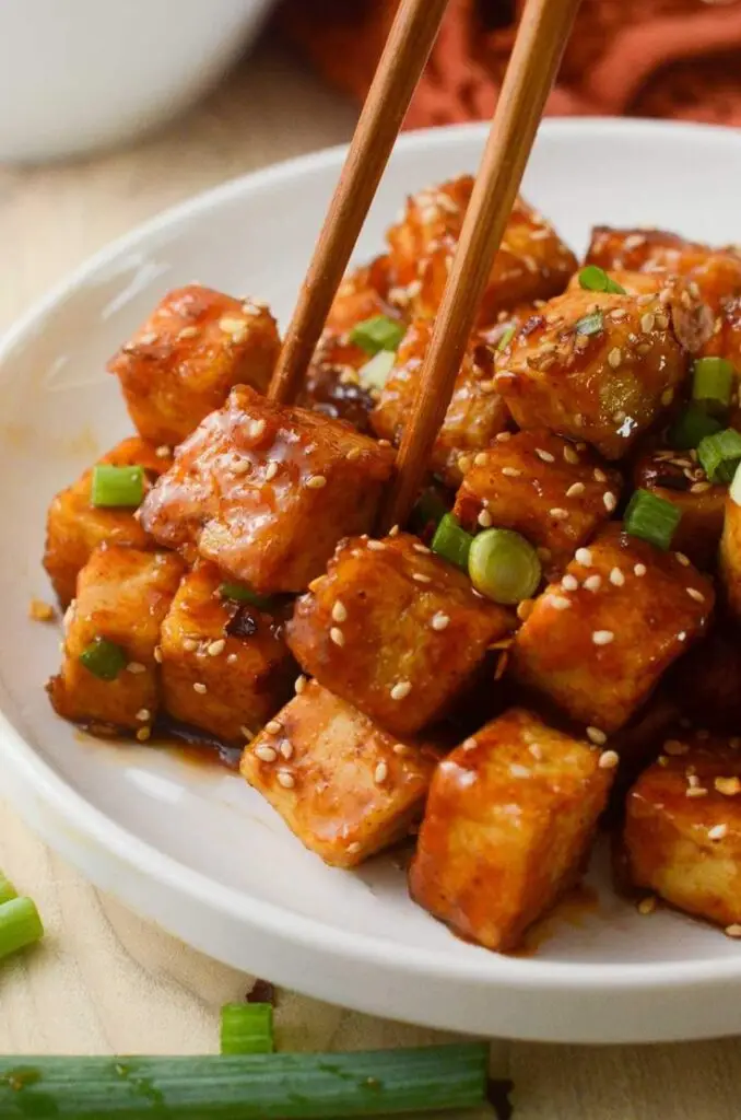 Spicy glazed tofu cubes garnished with sesame seeds and green onions, served on a white plate with chopsticks.