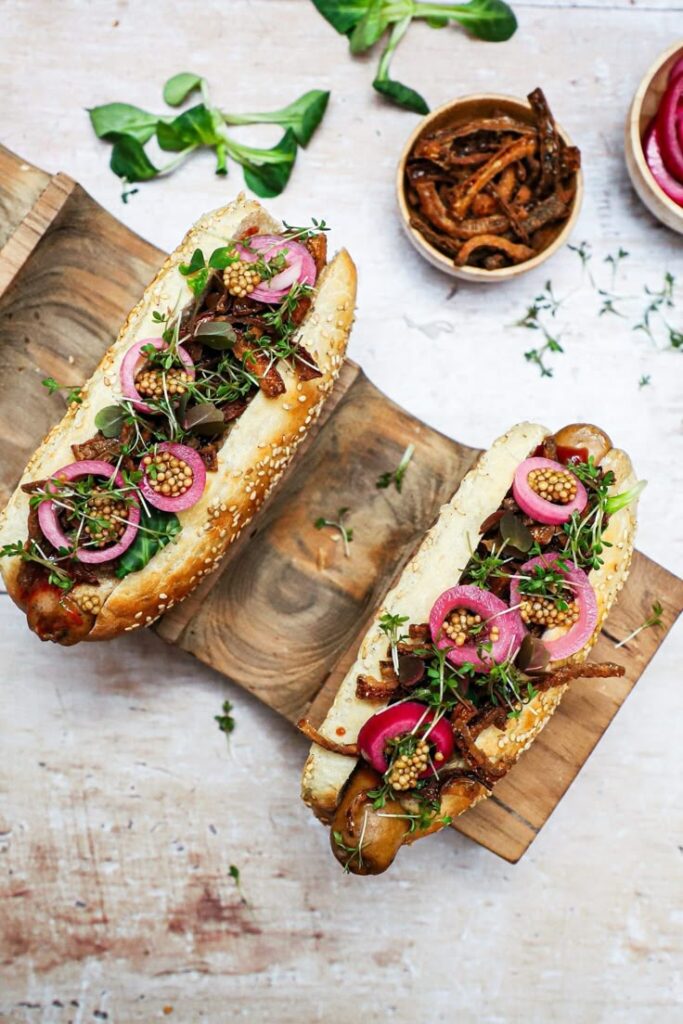 Gourmet hot dogs topped with pickled onions, mustard seeds, and herbs on a wooden board.