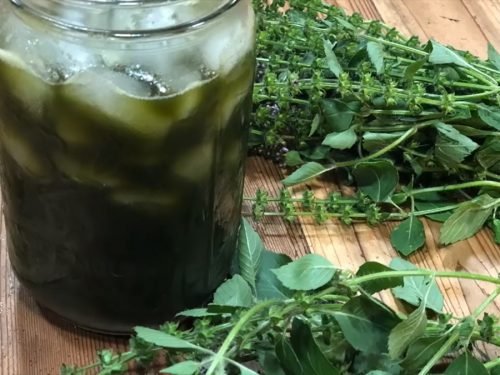 Jar of iced herbal drink next to fresh green herbs on a wooden table.