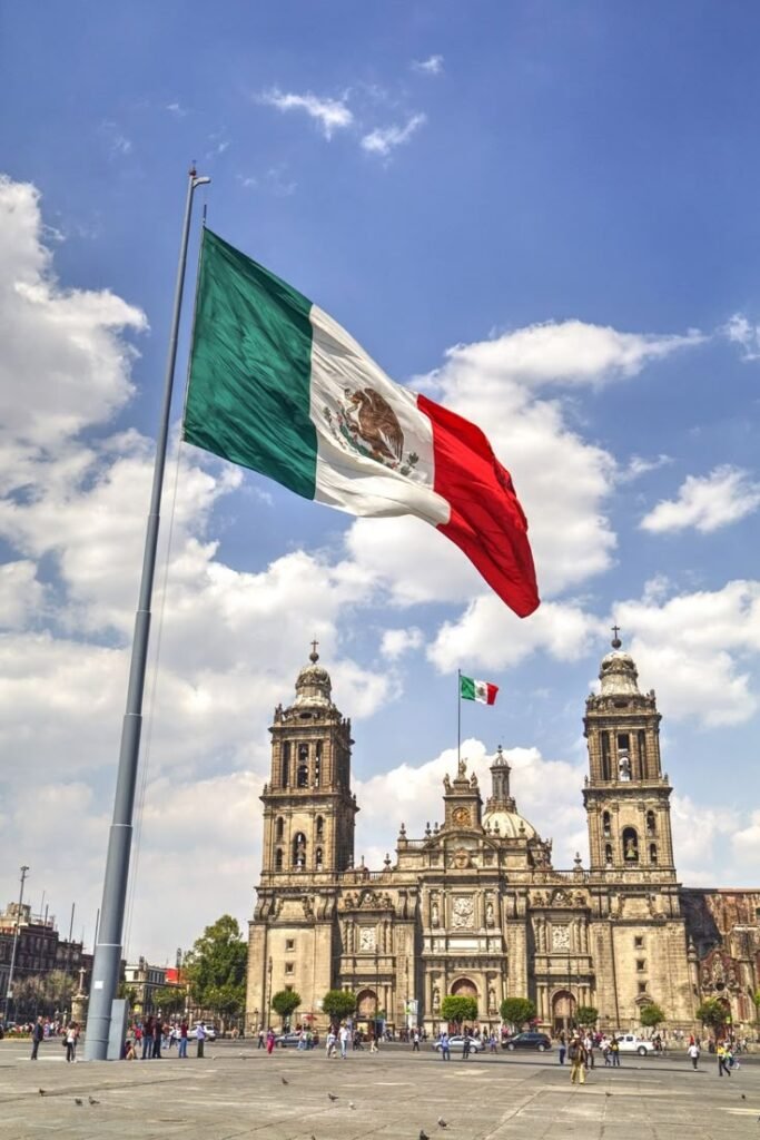 Mexican flag waving in front of the Metropolitan Cathedral in Mexico City under a bright blue sky.