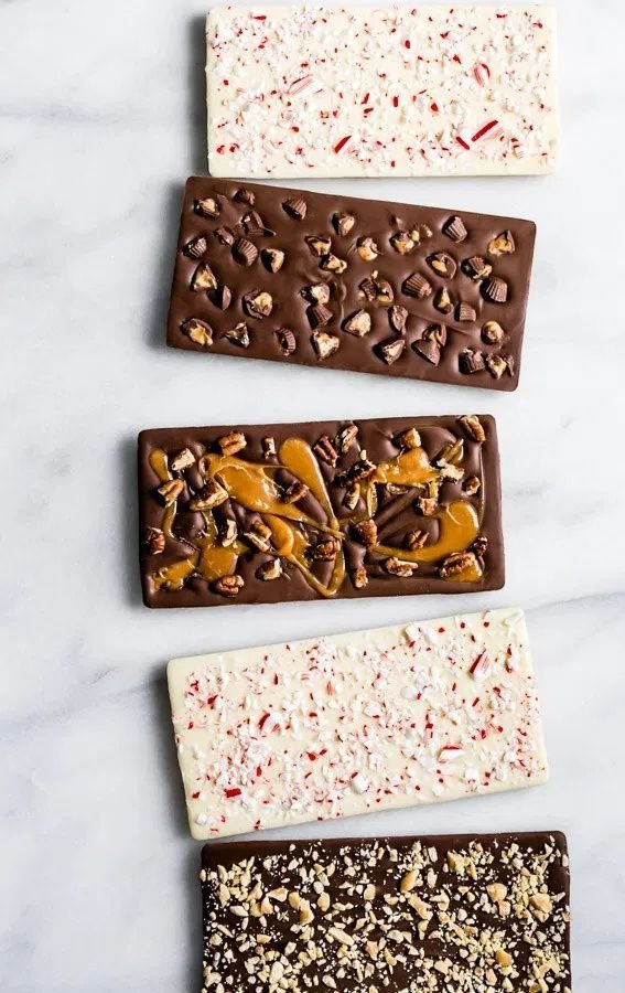 Five gourmet chocolate bars with assorted toppings on a marble surface.