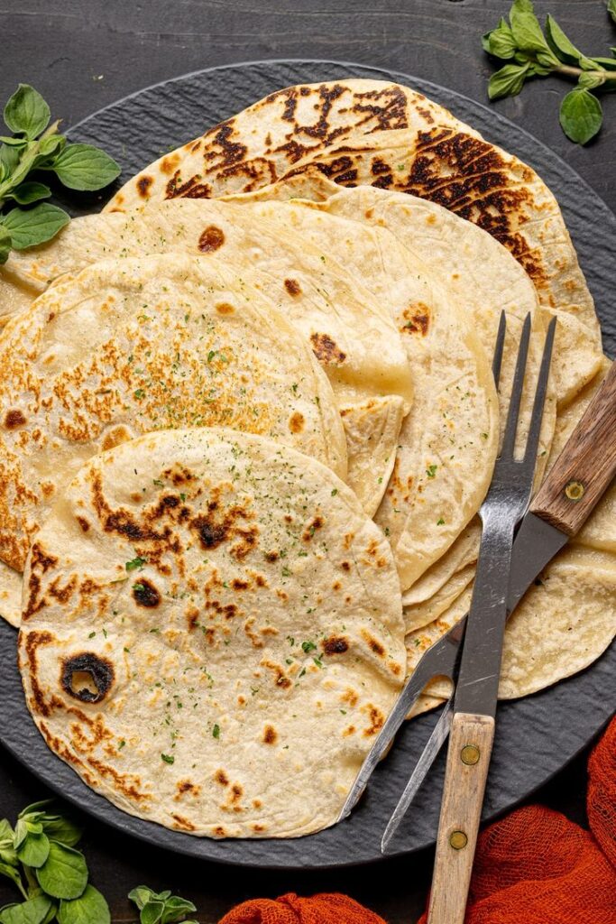 Fresh homemade flatbreads with herbs on a black plate, garnished with green herbs and wooden utensils.
