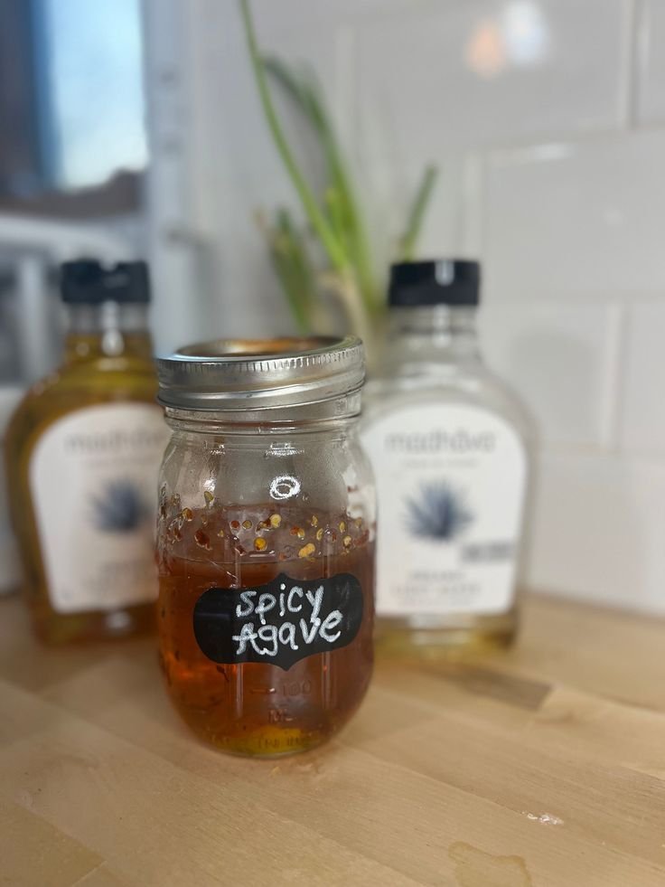 Jar of spicy agave syrup on a wooden table, with agave bottles blurred in the background.