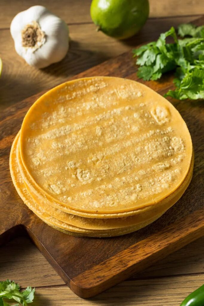 Fresh corn tortillas on a wooden board with cilantro, lime, and garlic on the side.