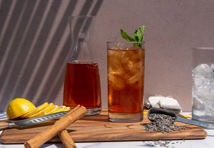 Iced tea with mint and lemon on a wooden board, surrounded by cinnamon sticks and lavender, in sunlight shadows.