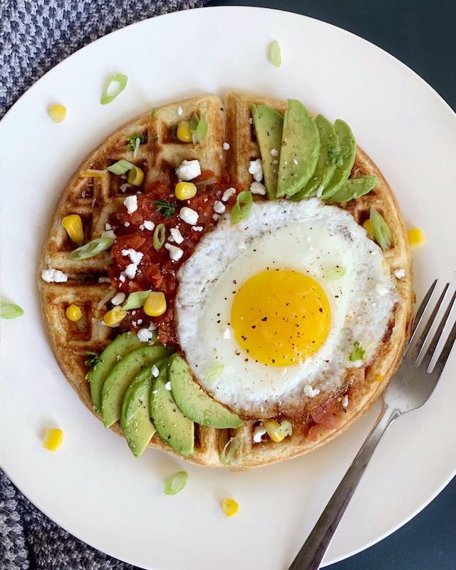 Waffle topped with avocado, salsa, corn, and a sunny-side-up egg, garnished with green onions and feta cheese.