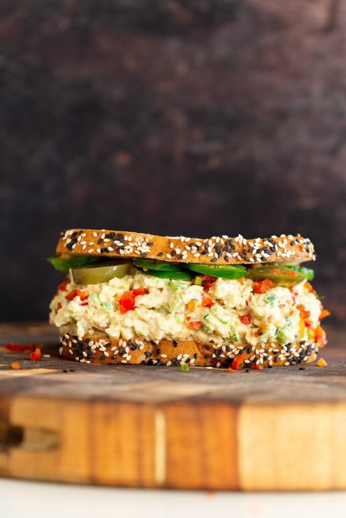 Delicious vegan chickpea salad sandwich on sesame seed bread with fresh vegetables, served on a rustic wooden board.