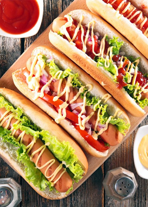 Delicious hot dogs topped with lettuce, onions, tomatoes, and sauces on a wooden board with condiments.